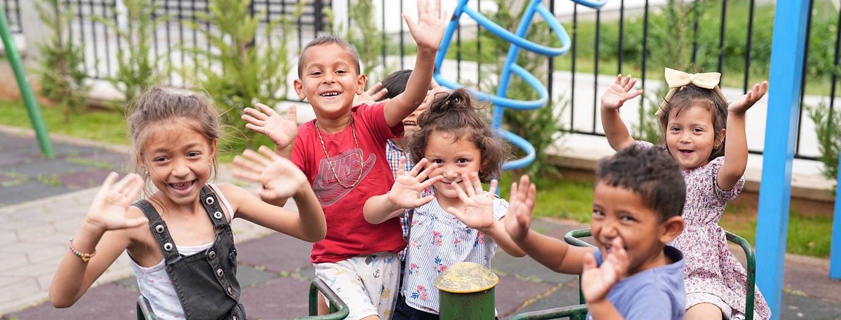 Children on a playground smiling and holding up their hands.