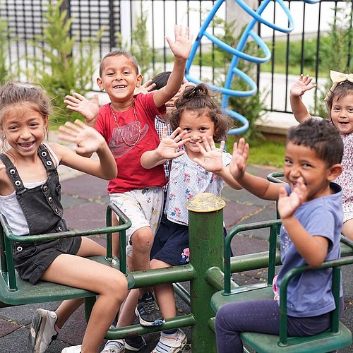 [Translate to English:] Kinder am Spielplatz lächeln und winken in die Kamera.