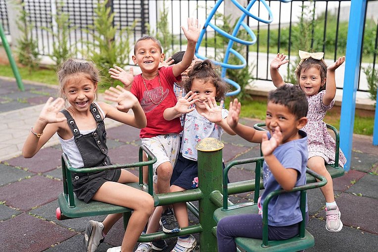 Children on a playground smiling and holding up their hands.